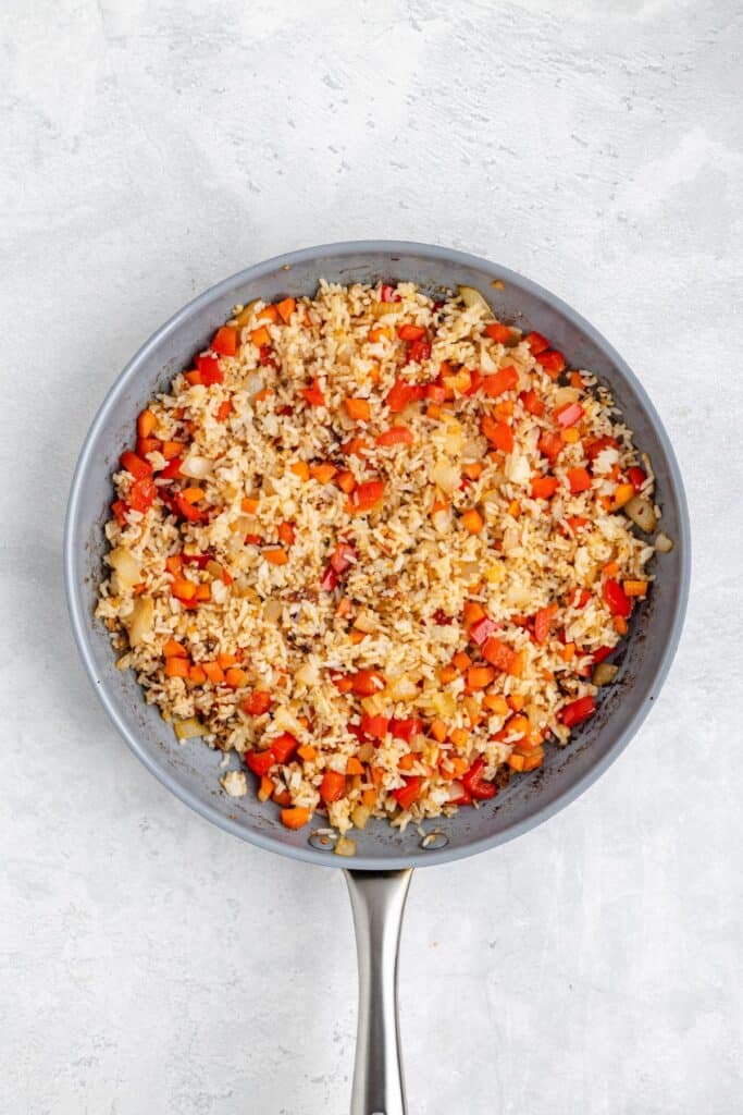 Frying pan with cooked rice, diced red bell peppers, onions, and mixed vegetables on a light gray surface.