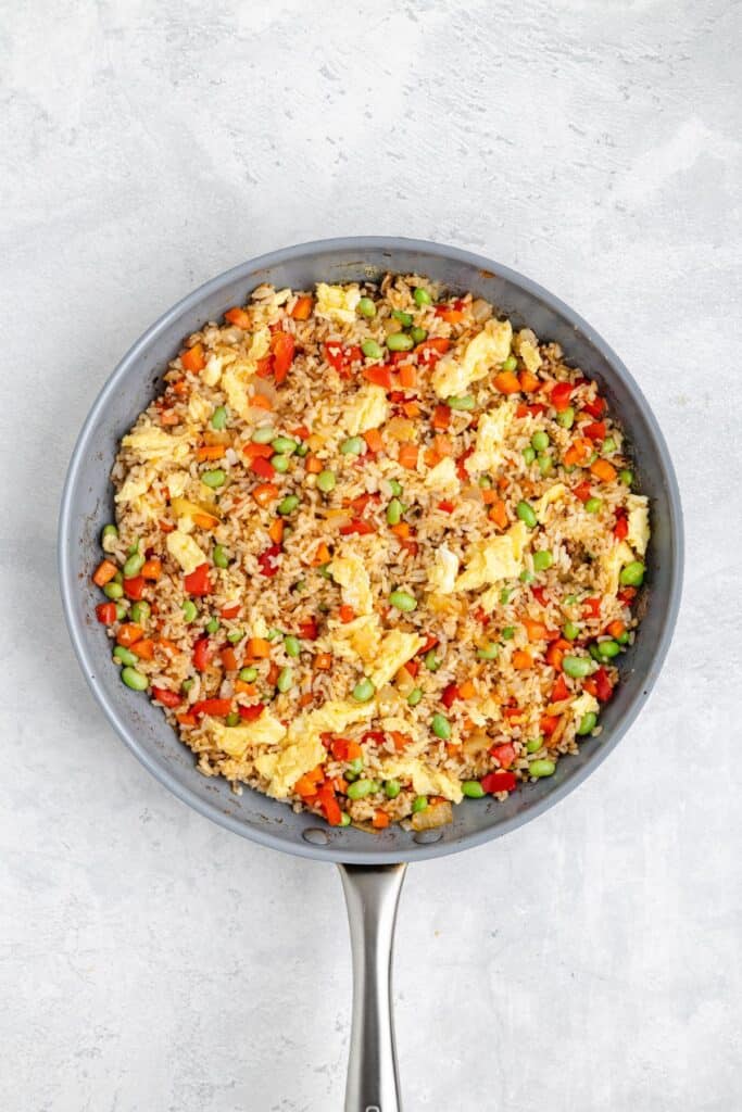 Frying pan of fried rice with eggs, peas, carrots, red bell peppers, and edamame on gray surface.