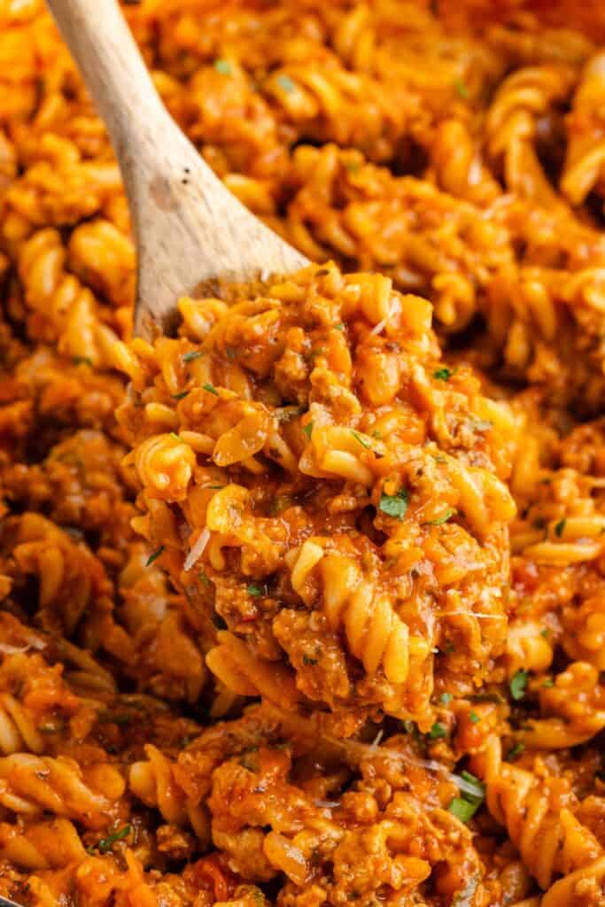 Wooden spoon lifting ground turkey and veggie pasta with herb garnish from a pan.