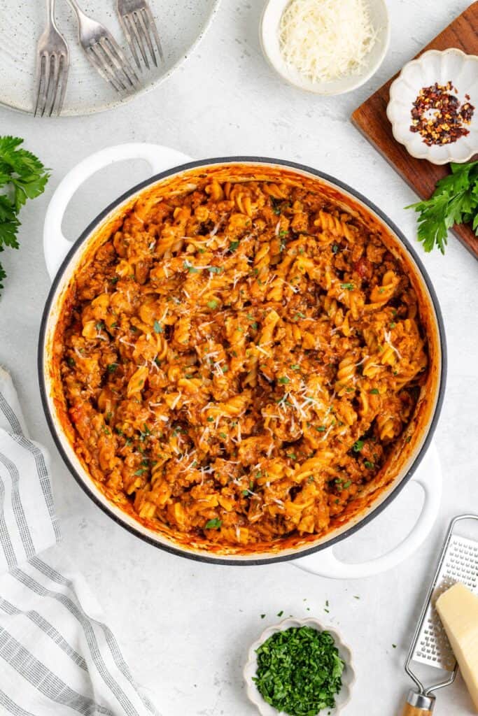 Large white pot of Ground Turkey Pasta with Zucchini topped with cheese and herbs, on light countertop with garnishes.