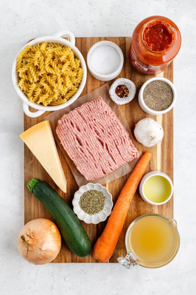 Ingredients for pasta dish: raw pasta, ground turkey, marinara sauce, cheese, chopped vegetables, oil, seasonings on board.