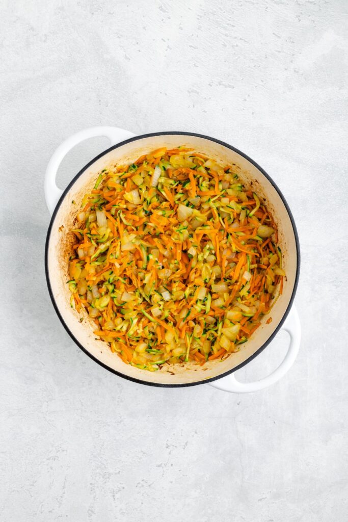 White pot with sautéed shredded carrots, zucchini, and onion on a light gray surface.