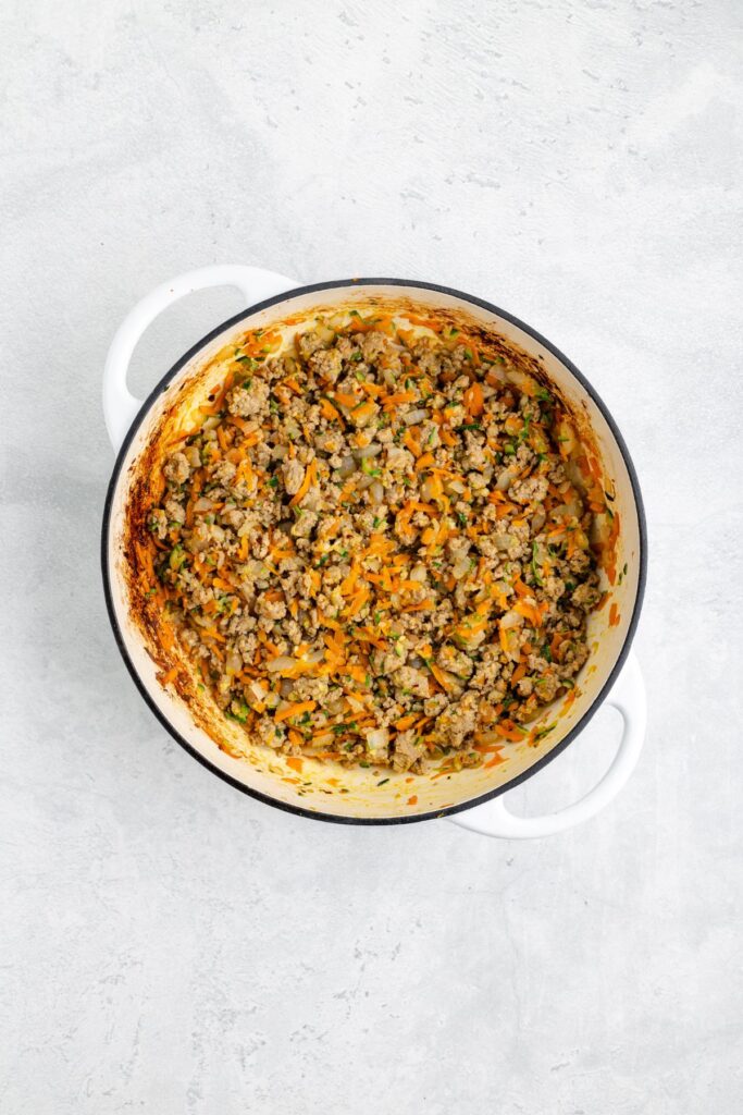 A white Dutch oven with cooked ground turkey, carrots, zucchini, onion, and herbs on a light gray background.