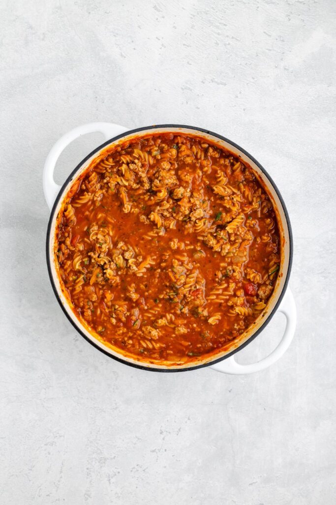 White pot with cooked ground turkey, pasta, and tomato sauce on a light gray surface.