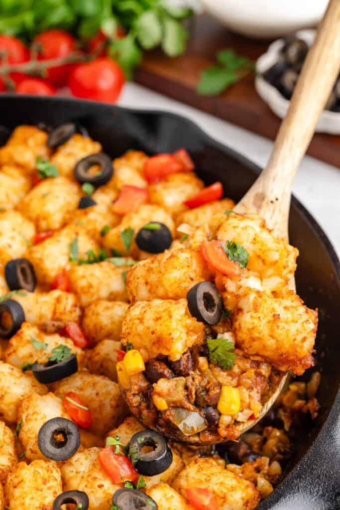 Wooden spoon lifting Mexican tater tot casserole with ground beef, corn, tomatoes, black olives, and herbs.