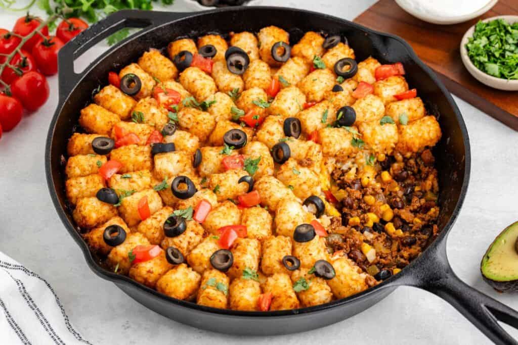 Cast iron skillet of taco tater tot casserole topped with tomatoes, black olives, herbs, ground meat, and corn showing.