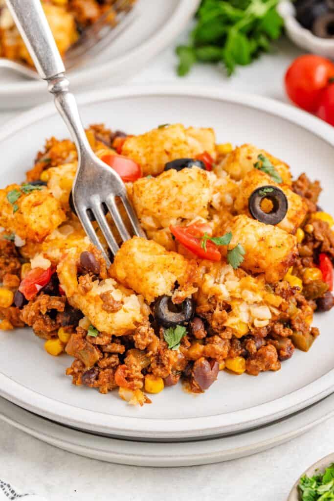 Plate of Mexican tater tot casserole with meat, beans, corn, tomatoes, cheese, olives, cilantro; fork resting on top.
