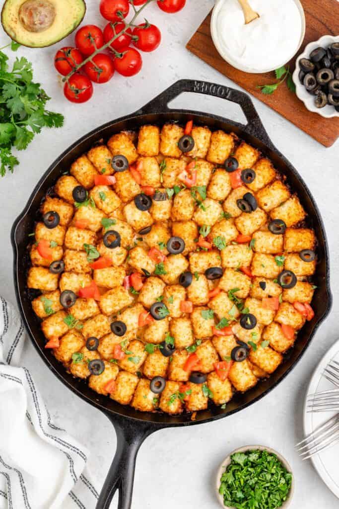 Cast iron skillet with cheesy tater tots, tomatoes, olives, and herbs, surrounded by fresh vegetables on white surface.