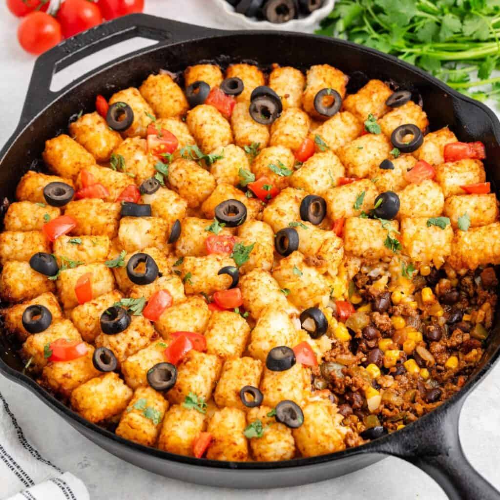 Baked tater tot casserole in cast iron skillet, topped with tomatoes, olives, herbs; beans and corn visible.