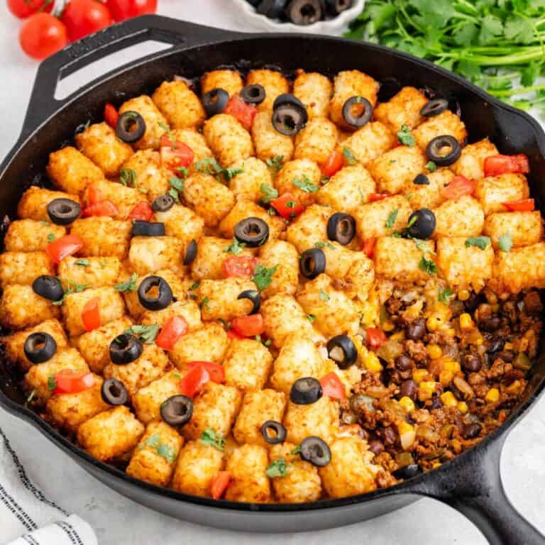 Mexican tater tot casserole in cast iron skillet, topped with tomatoes, olives, herbs; beans and corn visible.