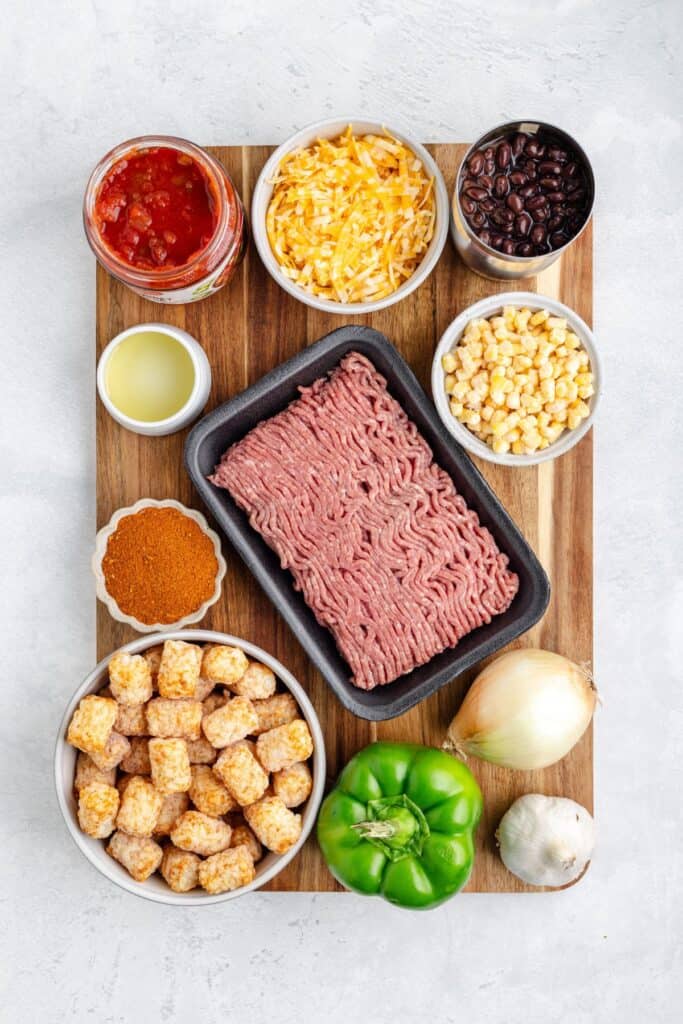Wooden board with bowls of ground meat, tater tots, cheese, salsa, beans, corn, peppers, and seasonings.