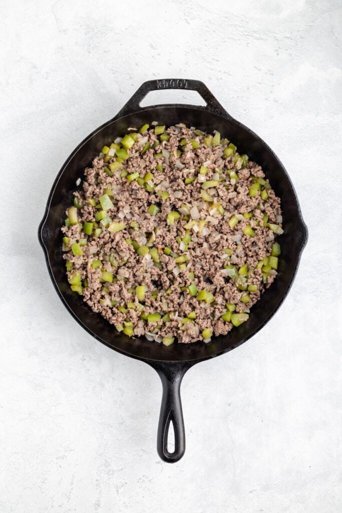 Cooked ground beef with chopped onions and peppers in a black cast iron skillet on a light surface.