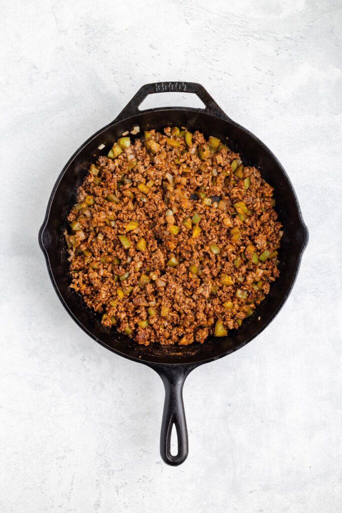 Cast iron skillet with cooked seasoned ground meat and diced vegetables on a light-colored surface.
