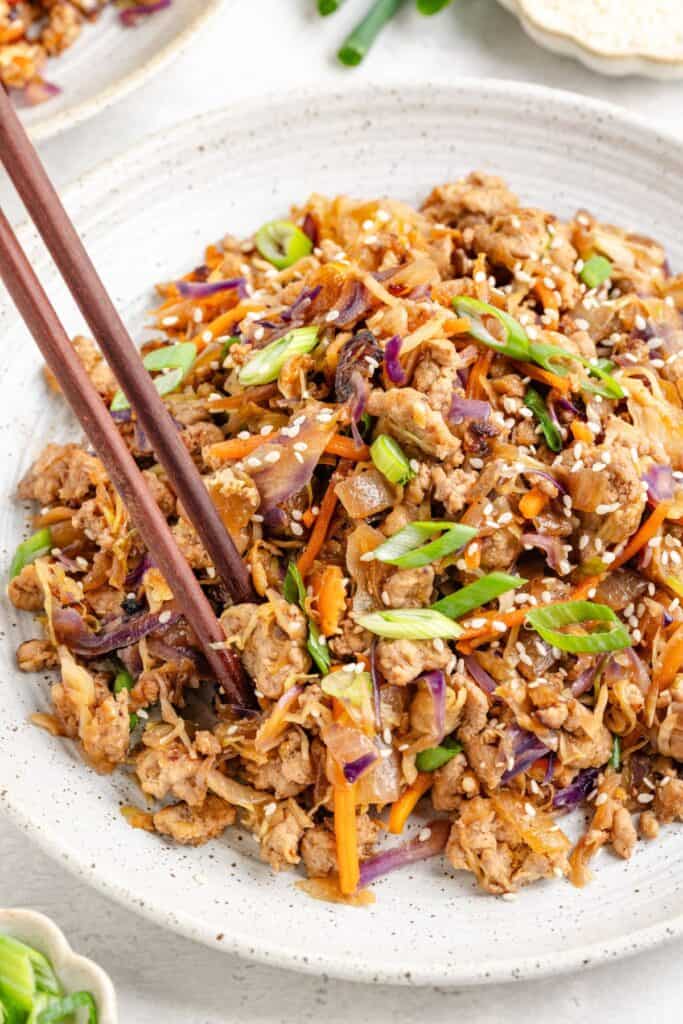 A bowl of ground meat stir-fry with carrots, cabbage, green onions, sesame seeds, and chopsticks.