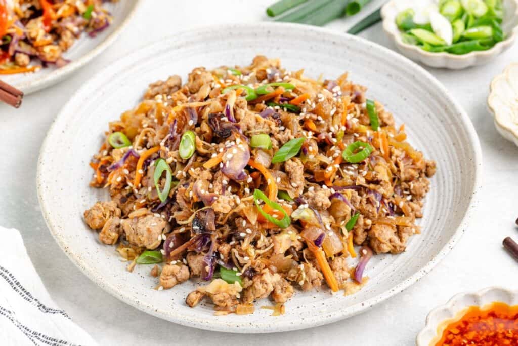 Pork egg roll in a bowl with shredded cabbage, carrots, mushrooms, green onions, and sesame seeds on a plate.