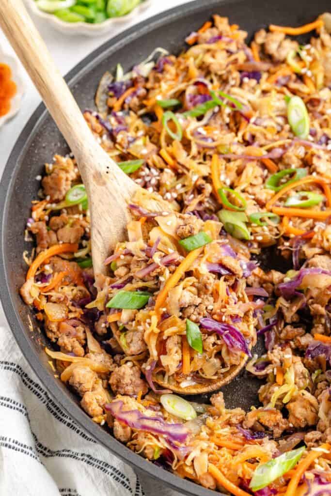 Skillet with Ground Pork Egg Roll in a Bowl, carrots, cabbage, and green onions being stirred with a wooden spoon.