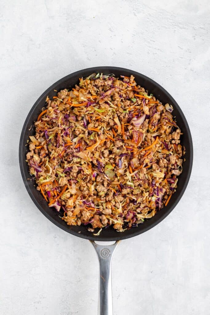 Skillet with cooked ground pork, shredded cabbage, carrots, and vegetables on a light gray background.