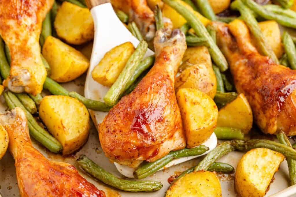 Baked BBQ chicken drumsticks, roasted potatoes, and green beans on a sheet pan with spatula lifting drumstick.