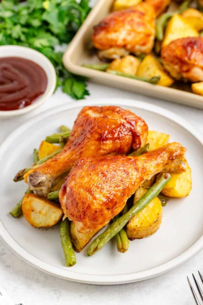 Two baked BBQ chicken drumsticks with roasted potatoes and green beans on a plate; barbecue sauce in background.