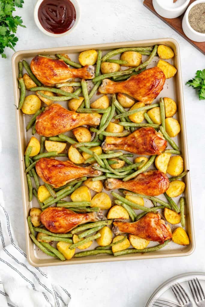 Sheet pan with baked BBQ chicken drumsticks, potatoes, and green beans; bowl of barbecue sauce on the side.