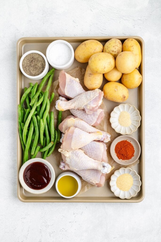 A baking tray with raw chicken drumsticks, green beans, baby potatoes, and small bowls of seasonings.