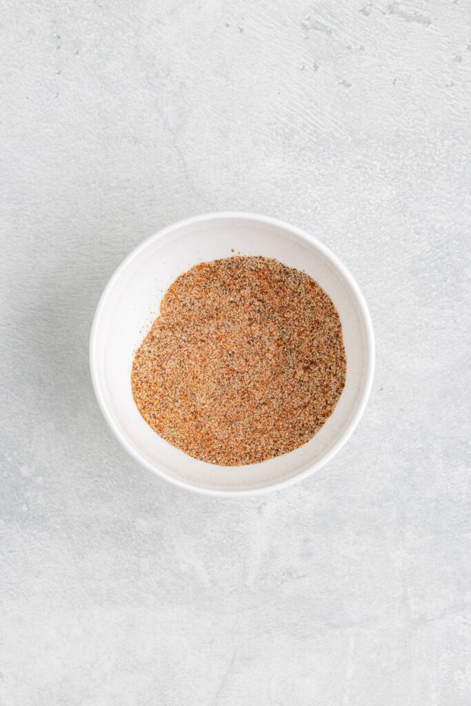 White bowl filled with BBQ spice blend on light gray textured background.