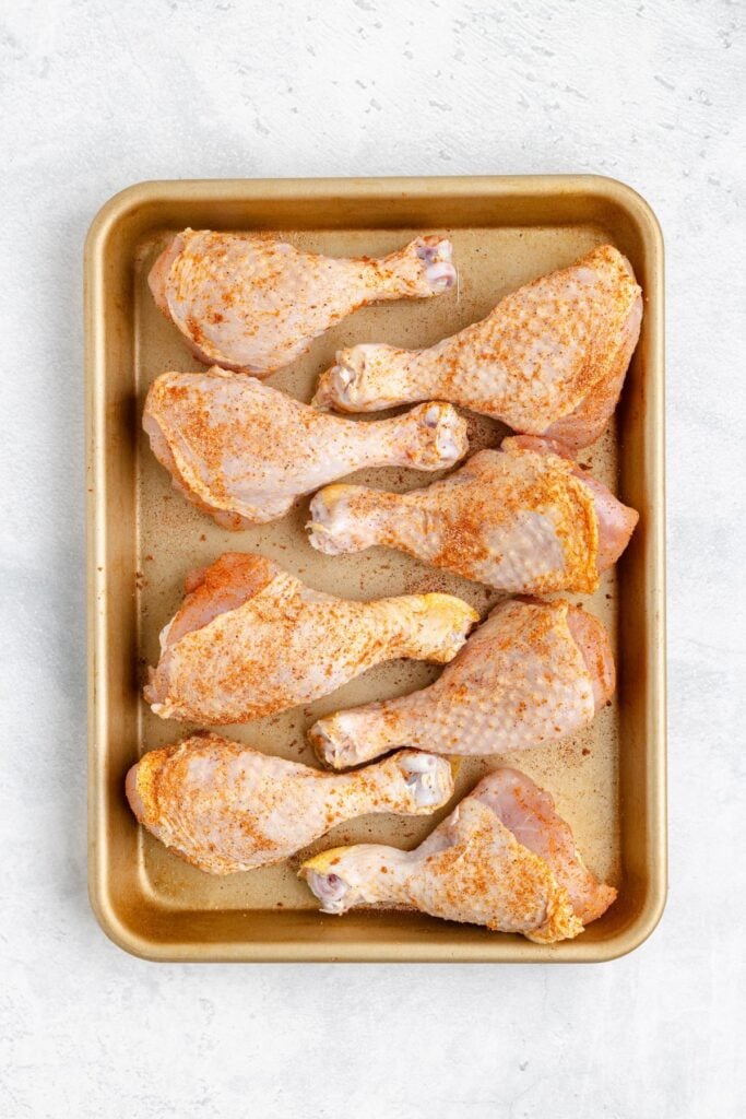 Baking tray with eight raw, seasoned chicken drumsticks arranged evenly on a light surface.
