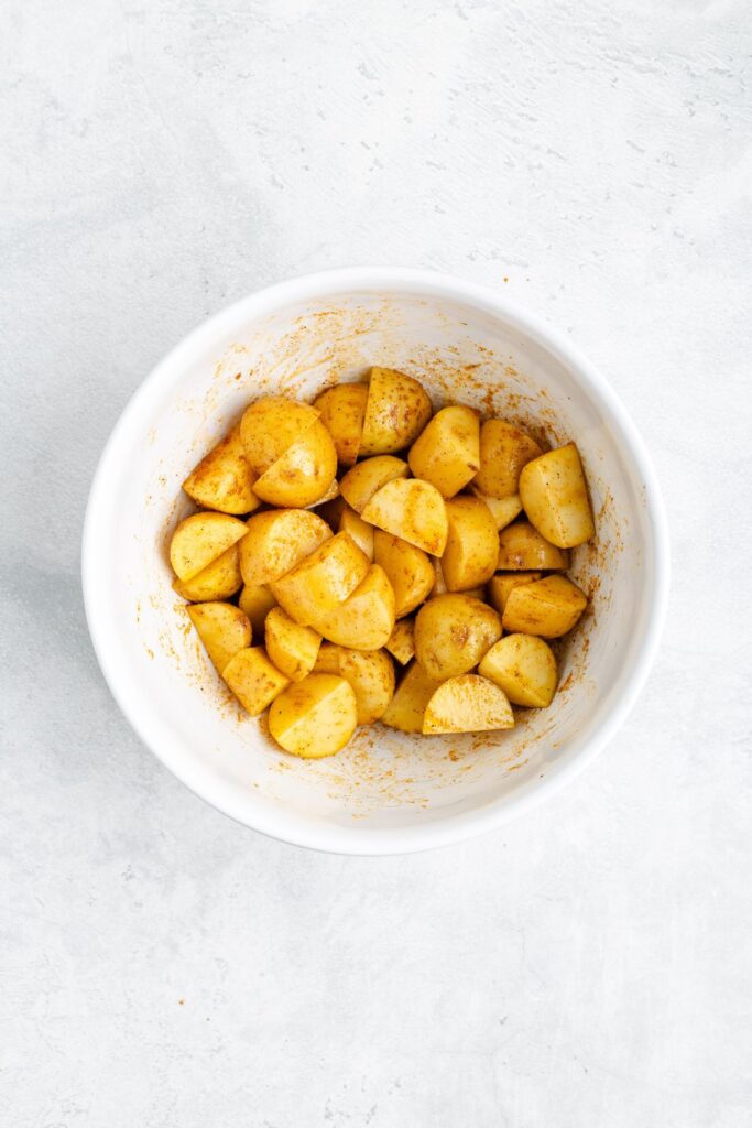 White bowl of spiced, quartered potatoes on a light surface.
