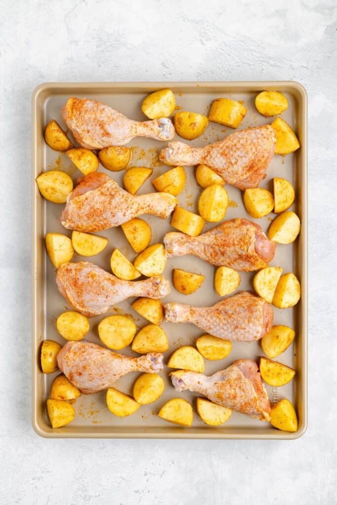 Raw chicken drumsticks and quartered yellow potatoes seasoned, arranged evenly on a baking sheet, ready to bake.