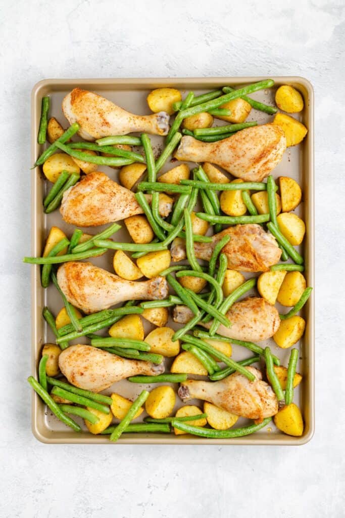 Par-cooked chicken drumsticks, halved potatoes, and green beans arranged on a baking sheet, ready for roasting.