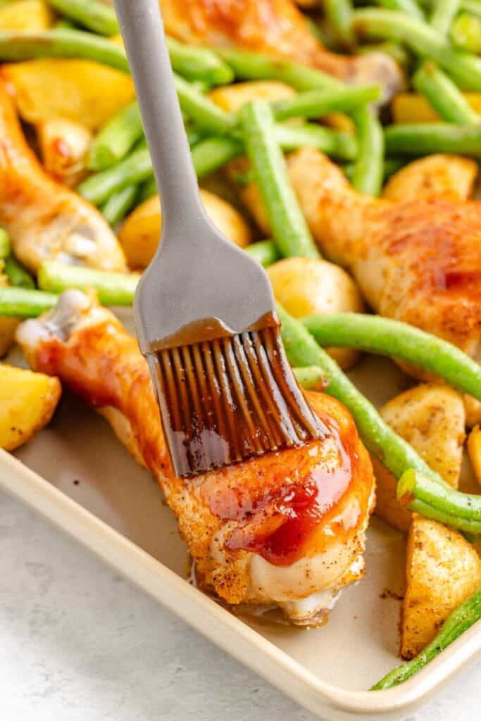 Silicone brush applying barbecue sauce to chicken drumstick with green beans and potato wedges on baking sheet.