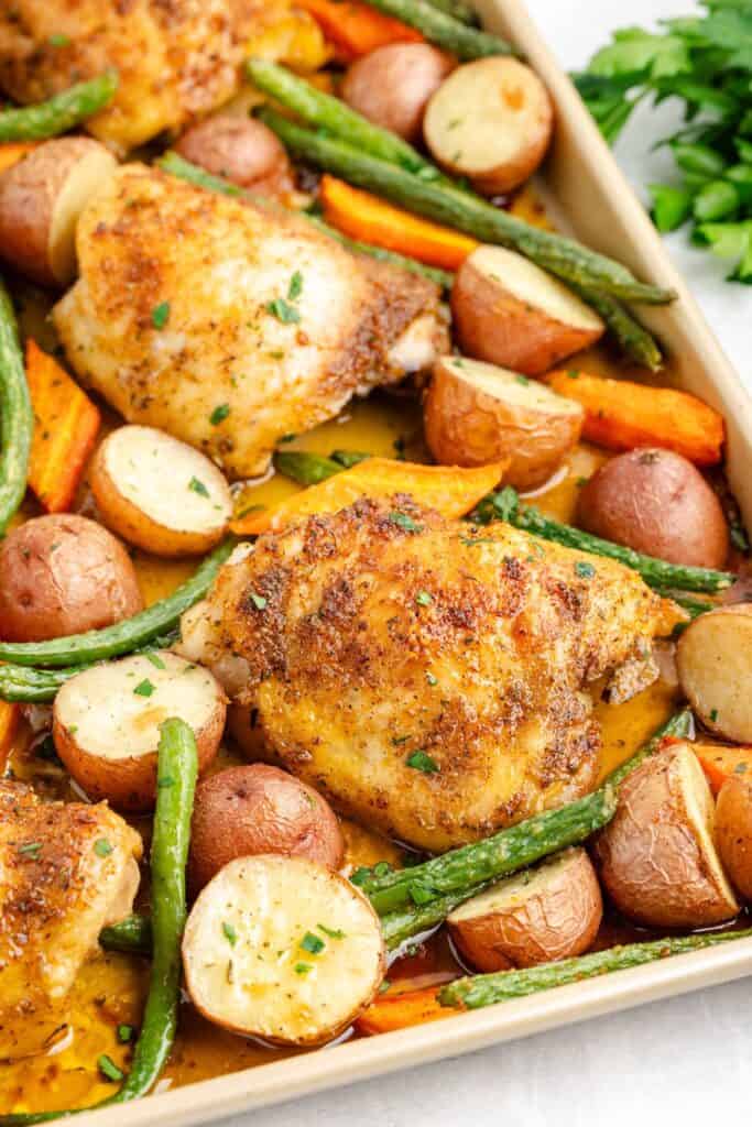 Sheet pan with baked chicken thighs, halved red potatoes, green beans, carrots, and fresh herb garnish.