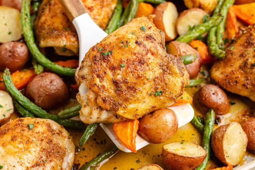 Sheet pan chicken thighs with golden skin on a spatula, surrounded by roasted potatoes, green beans, and carrots.