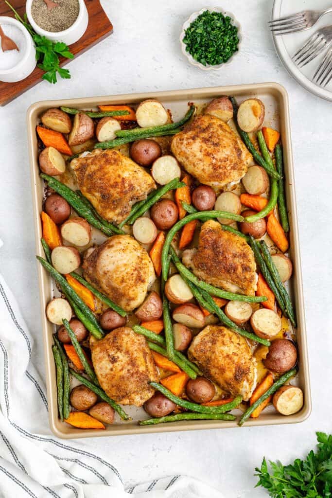 Sheet pan chicken thighs, potatoes, carrots, and green beans, surrounded by herbs and plates.