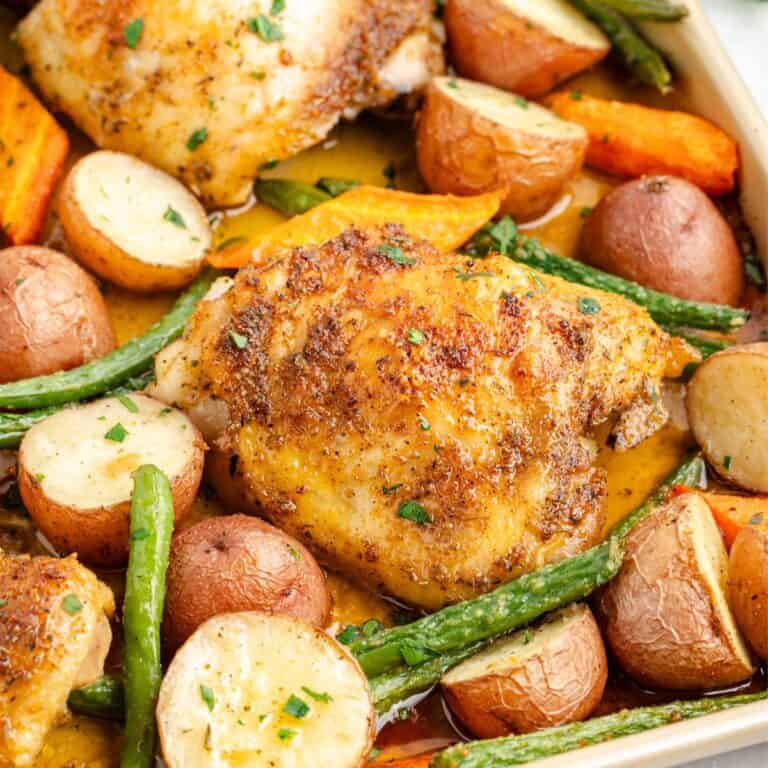 Baked chicken thighs and potatoes with green beans, and carrots, all herb-seasoned on a baking tray.