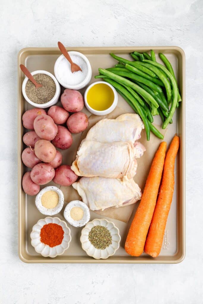 Raw chicken thighs, red potatoes, green beans, and carrots seasoned with spices on a baking tray before roasting.