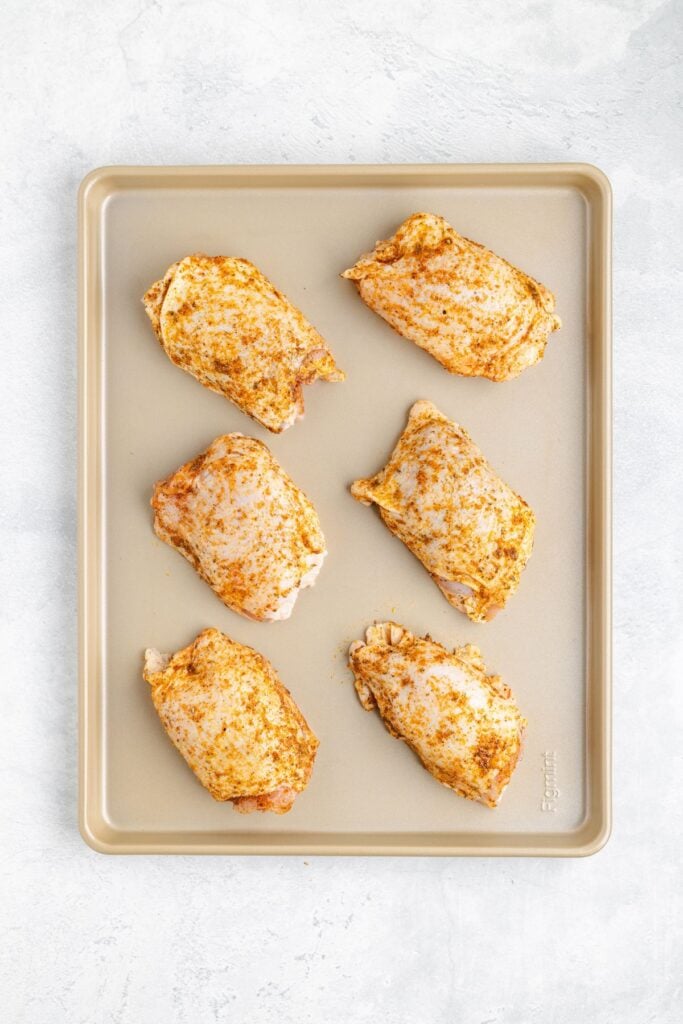 Six seasoned raw chicken thighs on a light baking sheet, overhead view, set on a white surface.