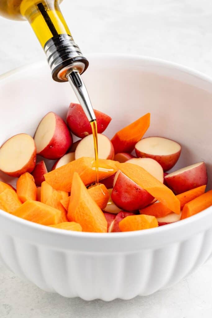 Olive oil pours over sliced carrots and halved red potatoes in a white bowl.