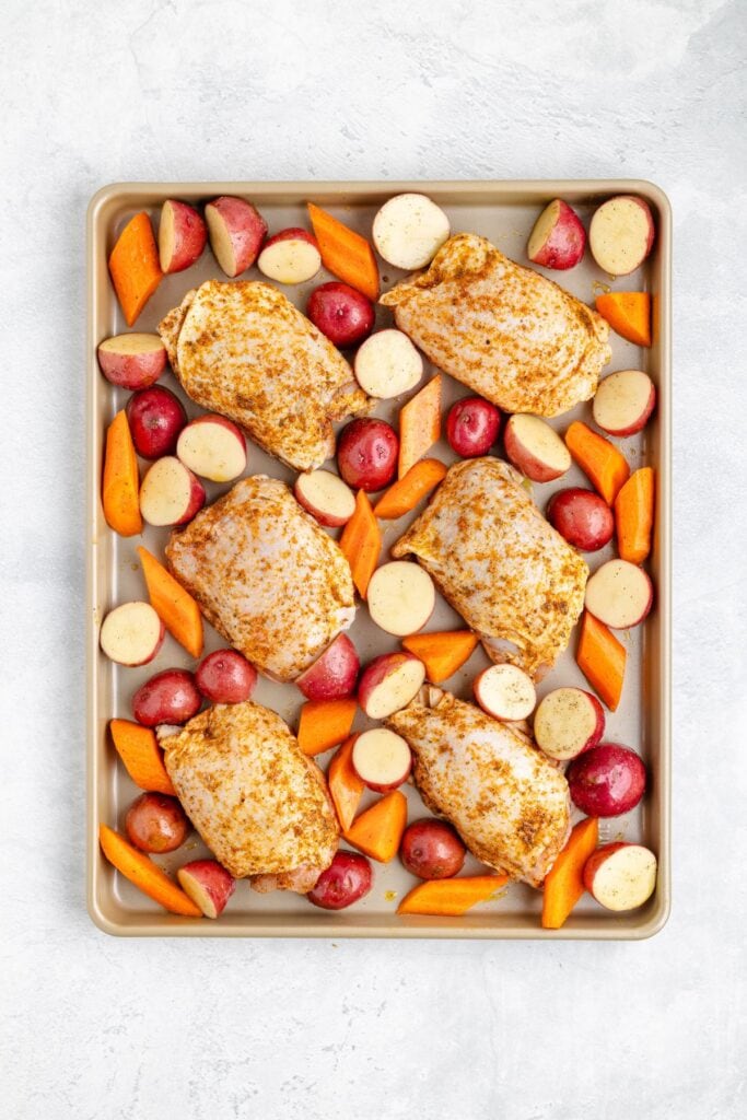 Raw chicken breasts, halved red potatoes, and sliced carrots arranged in rows on a baking sheet, uncooked.