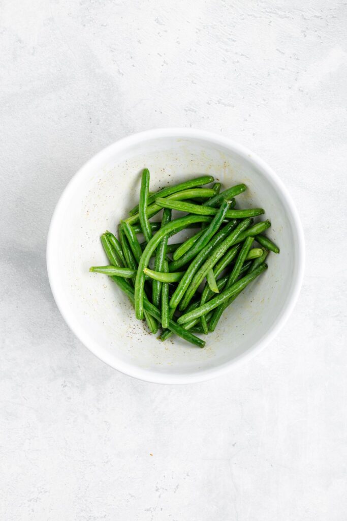 White bowl of fresh green beans seasoned with black pepper on a light gray surface.