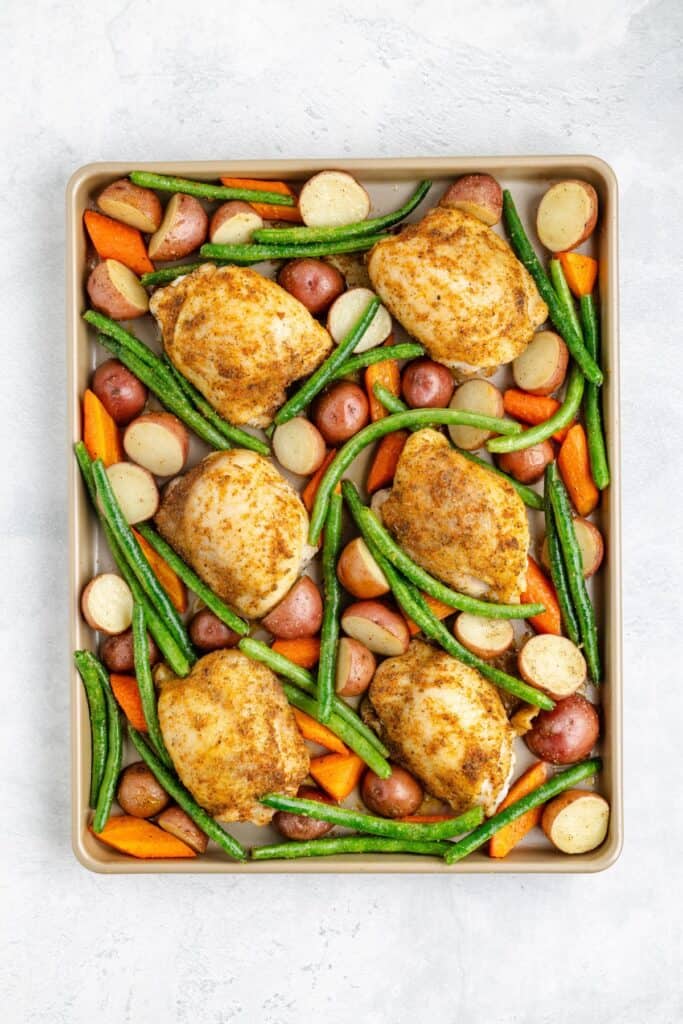 Sheet pan with six seasoned chicken thighs, halved baby potatoes, green beans, and sliced carrots on light background.