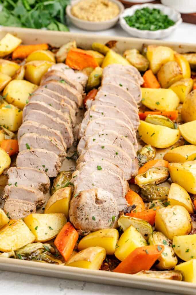 Sliced roasted pork tenderloin with potatoes, carrots, Brussels sprouts, and chopped herbs on a sheet pan.