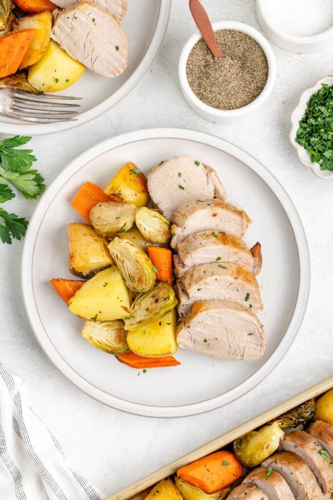 Plate of sliced pork tenderloin with roasted potatoes, carrots, Brussels sprouts, and herbs; fork and seasonings beside.