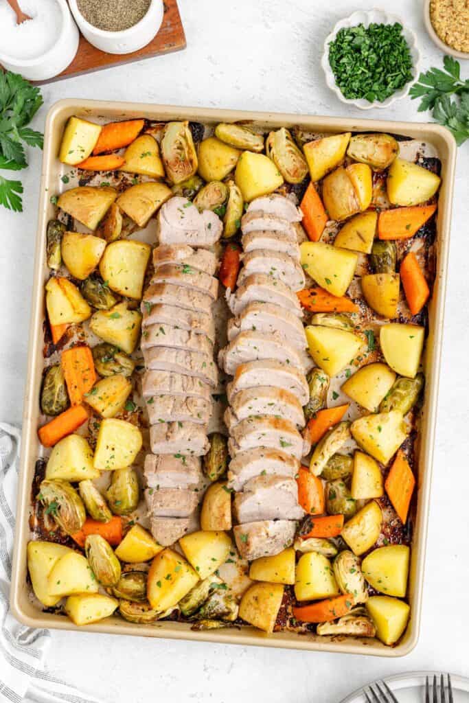 Two sliced pork tenderloins on a sheet pan with roasted potatoes, carrots, and Brussels sprouts.