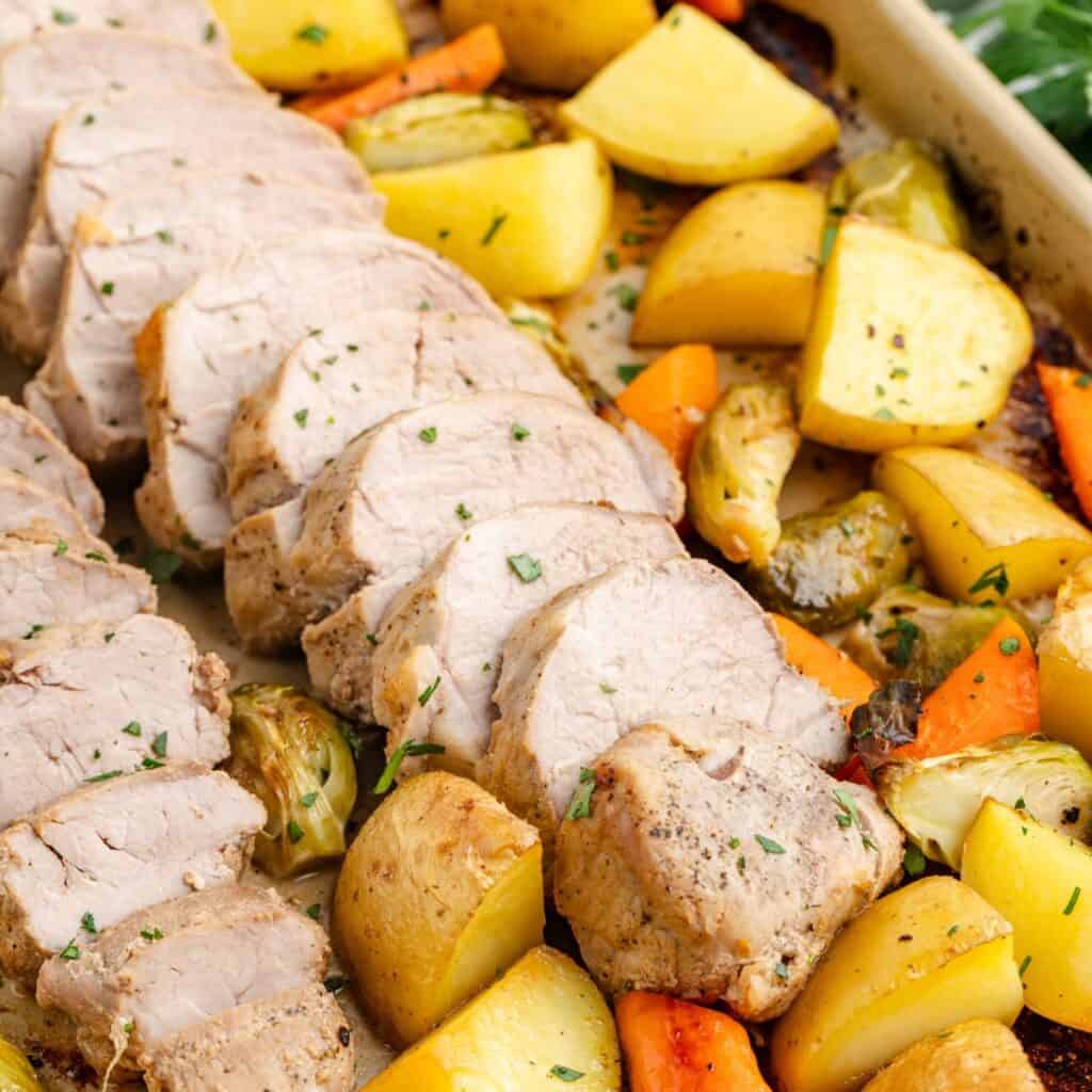 Sliced pork tenderloin with roasted potatoes, carrots, and Brussels sprouts on a sheet pan.