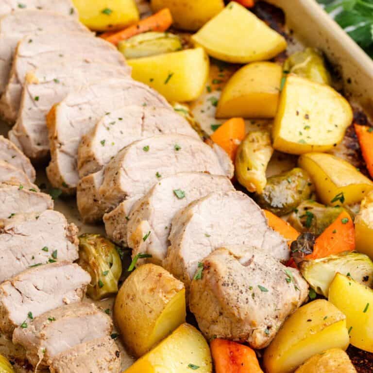 Sliced pork tenderloin with roasted potatoes, carrots, and Brussels sprouts on a sheet pan.