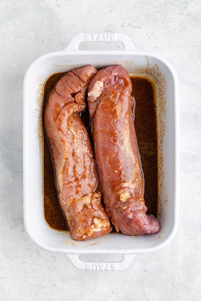 Two raw pork tenderloins marinating in brown sauce inside a rectangular white baking dish.