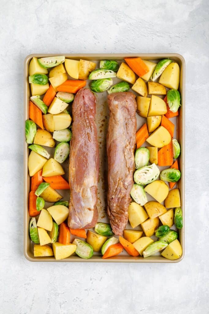 Two raw pork tenderloins on a baking sheet with chopped potatoes, carrots, and Brussels sprouts, ready to roast.