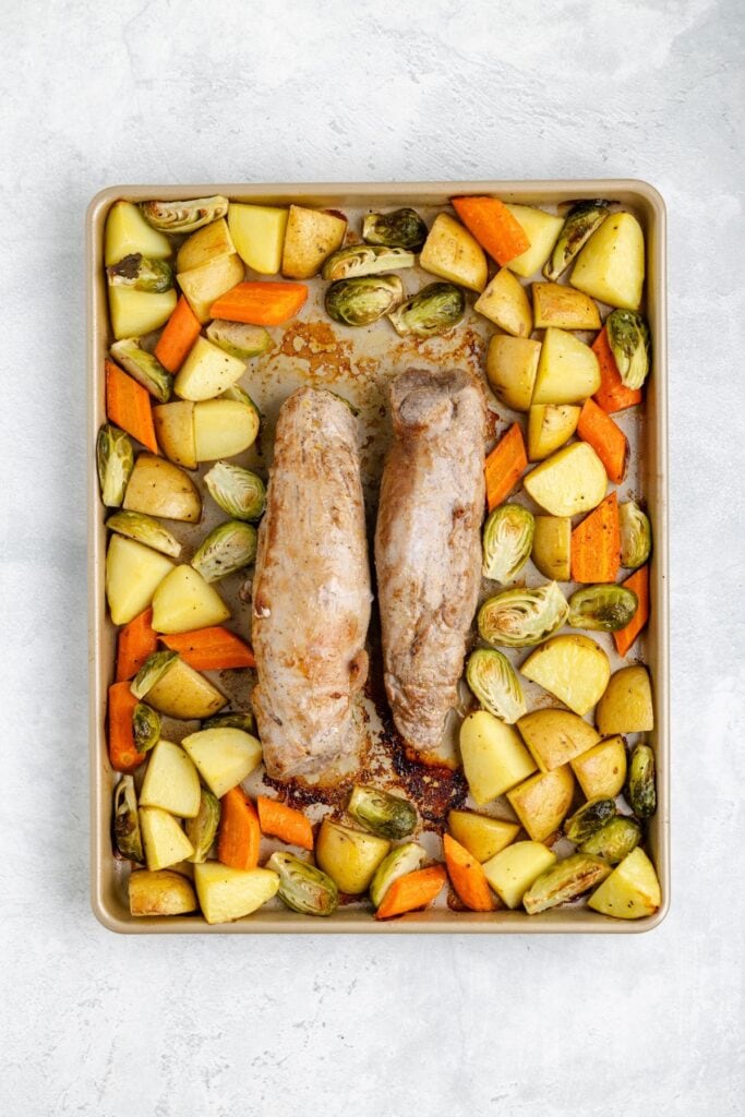 Two pork tenderloins on a baking sheet, surrounded by roasted potatoes, carrots, and halved Brussels sprouts.