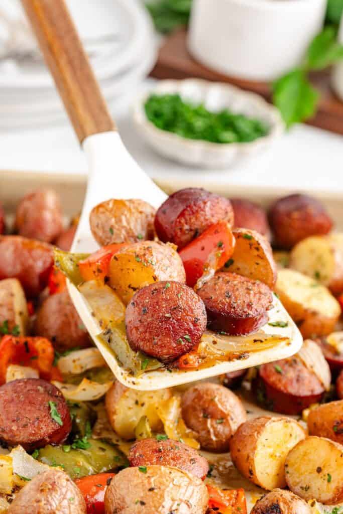 A spatula lifts roasted sausage, potatoes, and vegetables from a sheet pan; fresh herbs are visible behind.